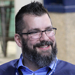 a person with glasses, short brown hair and a beard smiles in a close up shot.