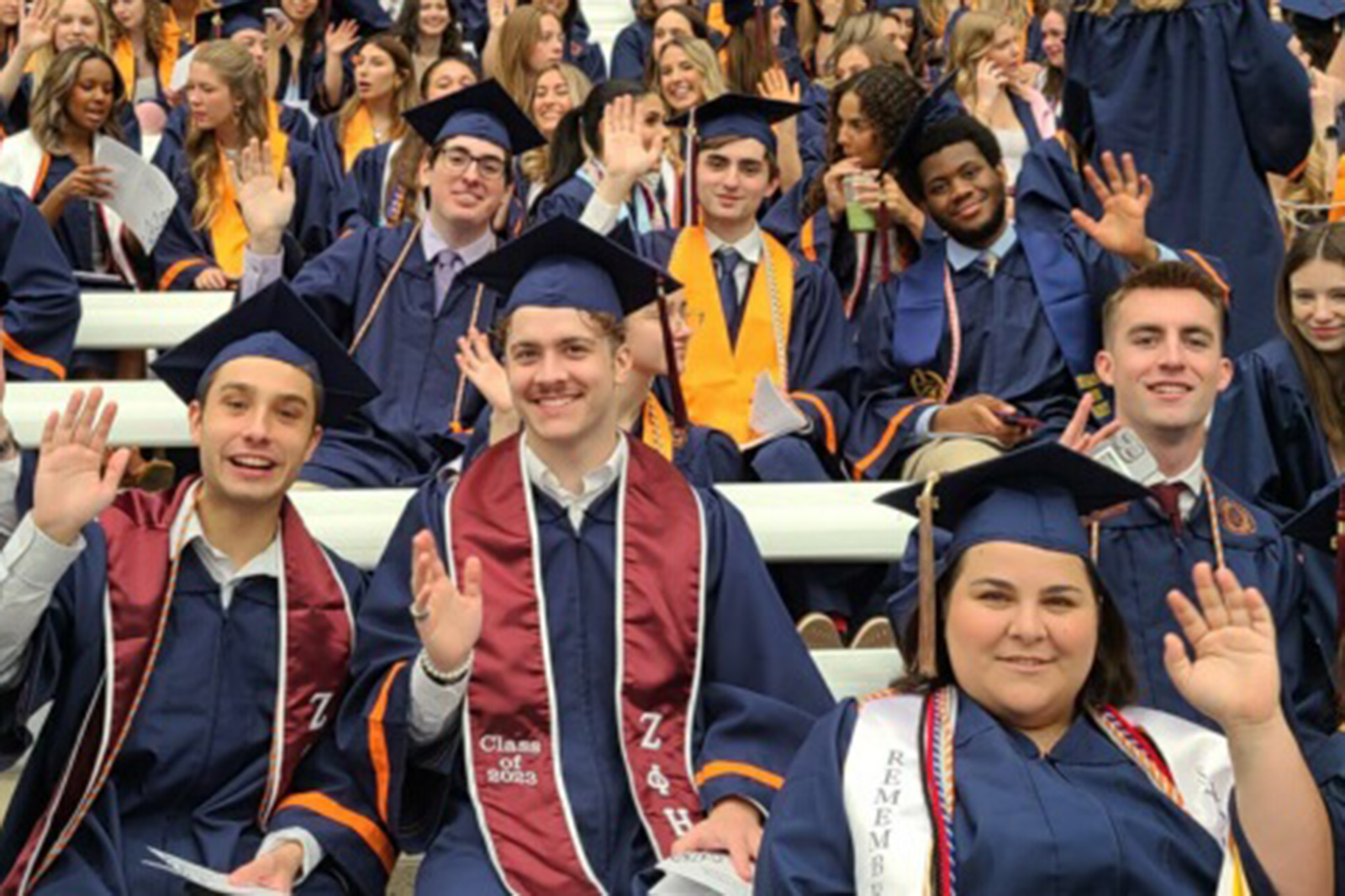 Students in academic regalia wave during a photo while sitting on bleachers