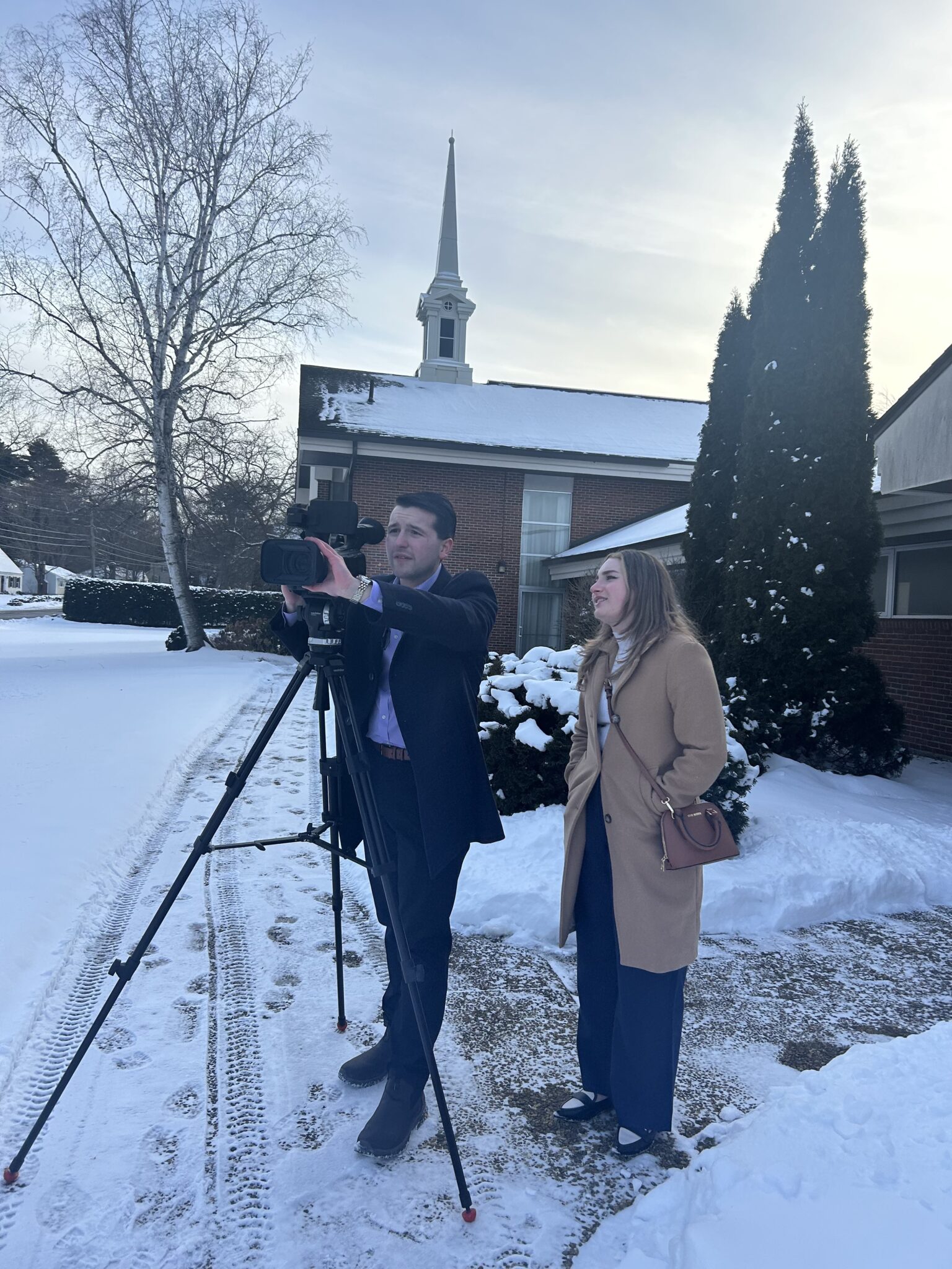 two students adjust a news camera while standing outside in the snow