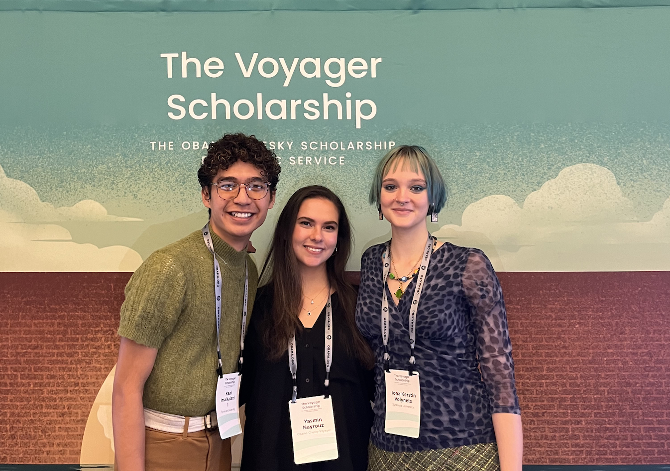 3 people stand and smile together under the voyager scholarship sign