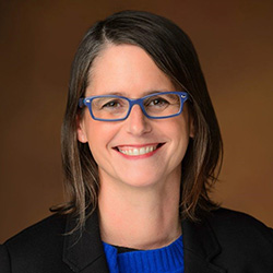 a person with medium length brown hair and blue glasses wears a blue shirt and black blazer while posing for a headshot