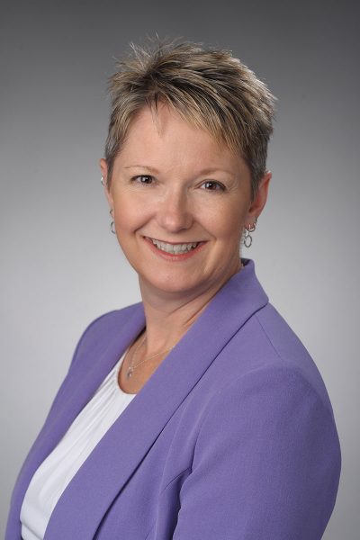 a person with short blonde hair and earrings wears a white shirt and purple blazer while posing for a headshot