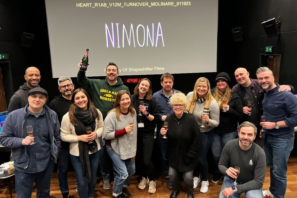 a group of people stand in front of a screen that says "Nimona" and hold champagne flutes