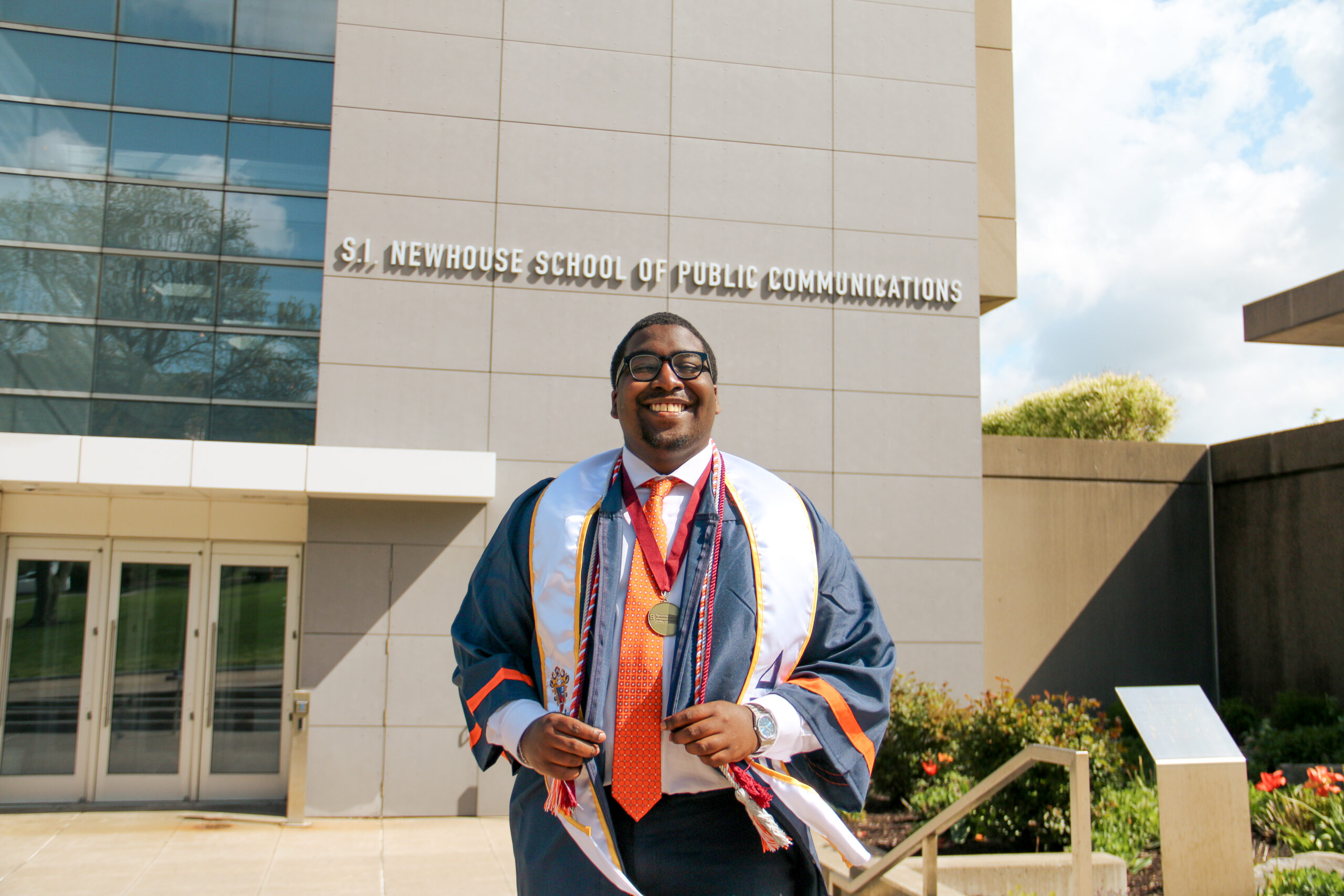 a person in a graduation gown stands outside and smiles