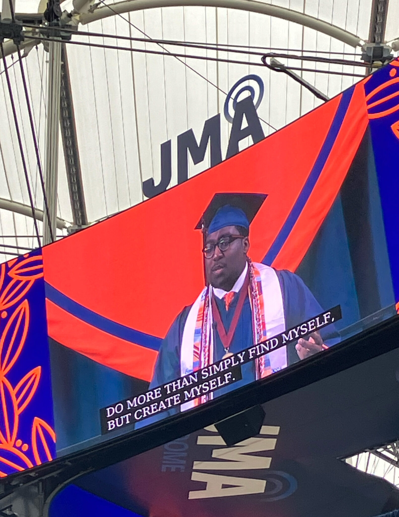 A man is shown on a video board speaking during a convocation ceremony