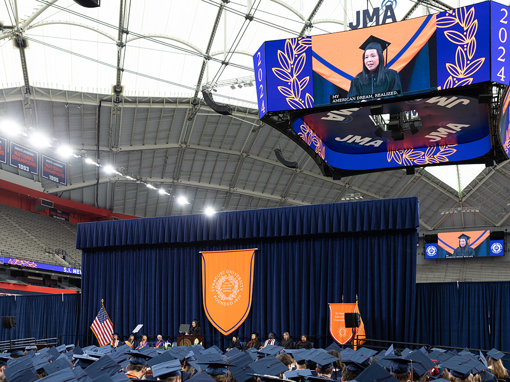 A woman is shown speaking on a video screen above a stage during a convocation ceremony
