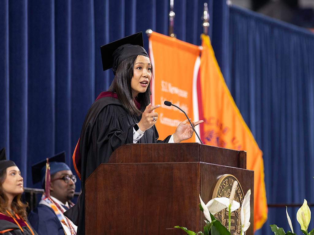 A woman dressed in a graduation cap and gown speaks at a podium during a convocation ceremony