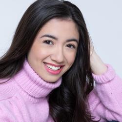 person with long dark hair and dark eyes wears a pink turtleneck and poses for a headshor