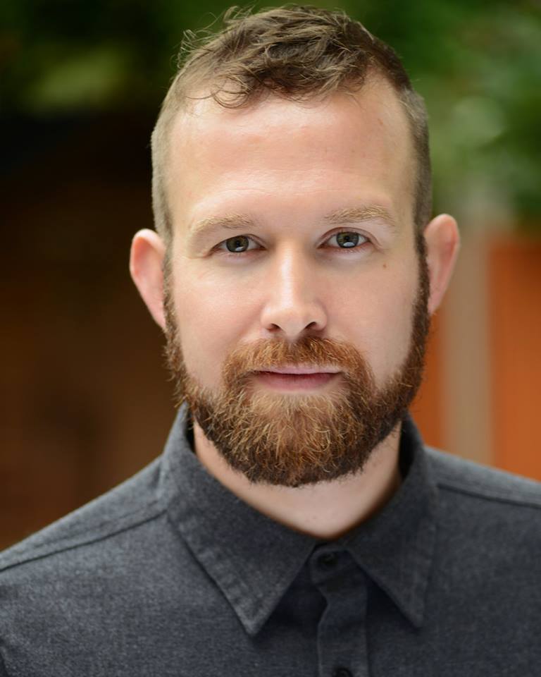 a person with light brown short hair and a beard poses for a headshot