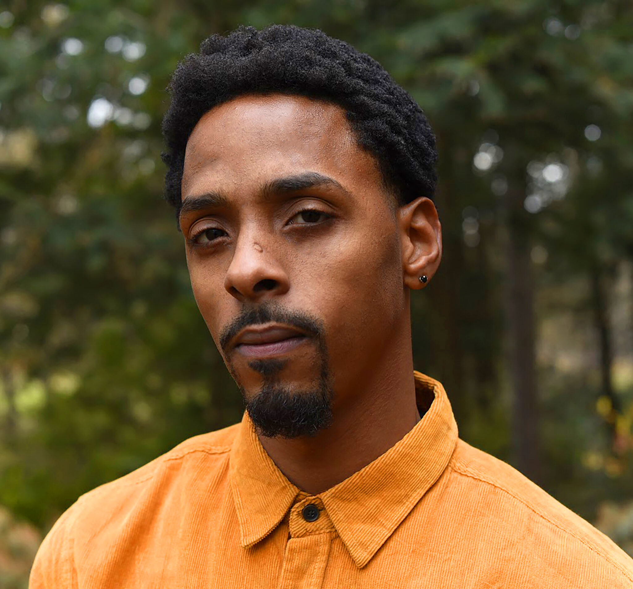 person with short dark hair and facial hair wears an orange shirt and poses for a headshot