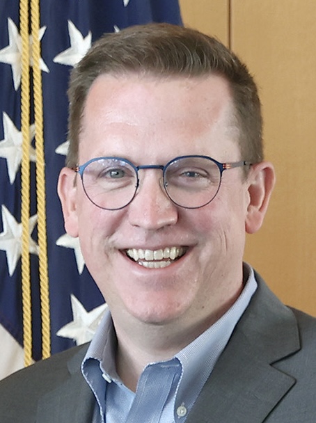 a person with glasses and short brown hair poses for a headshot in front of the American Flag