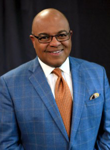 a person with a bald head and glasses poses for a headshot in a blue plaid balzer, white shirt and orange tie