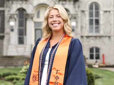 a person with blonde hair smiles for the camera as they pose outside in a graduation gown and tassels around their neck