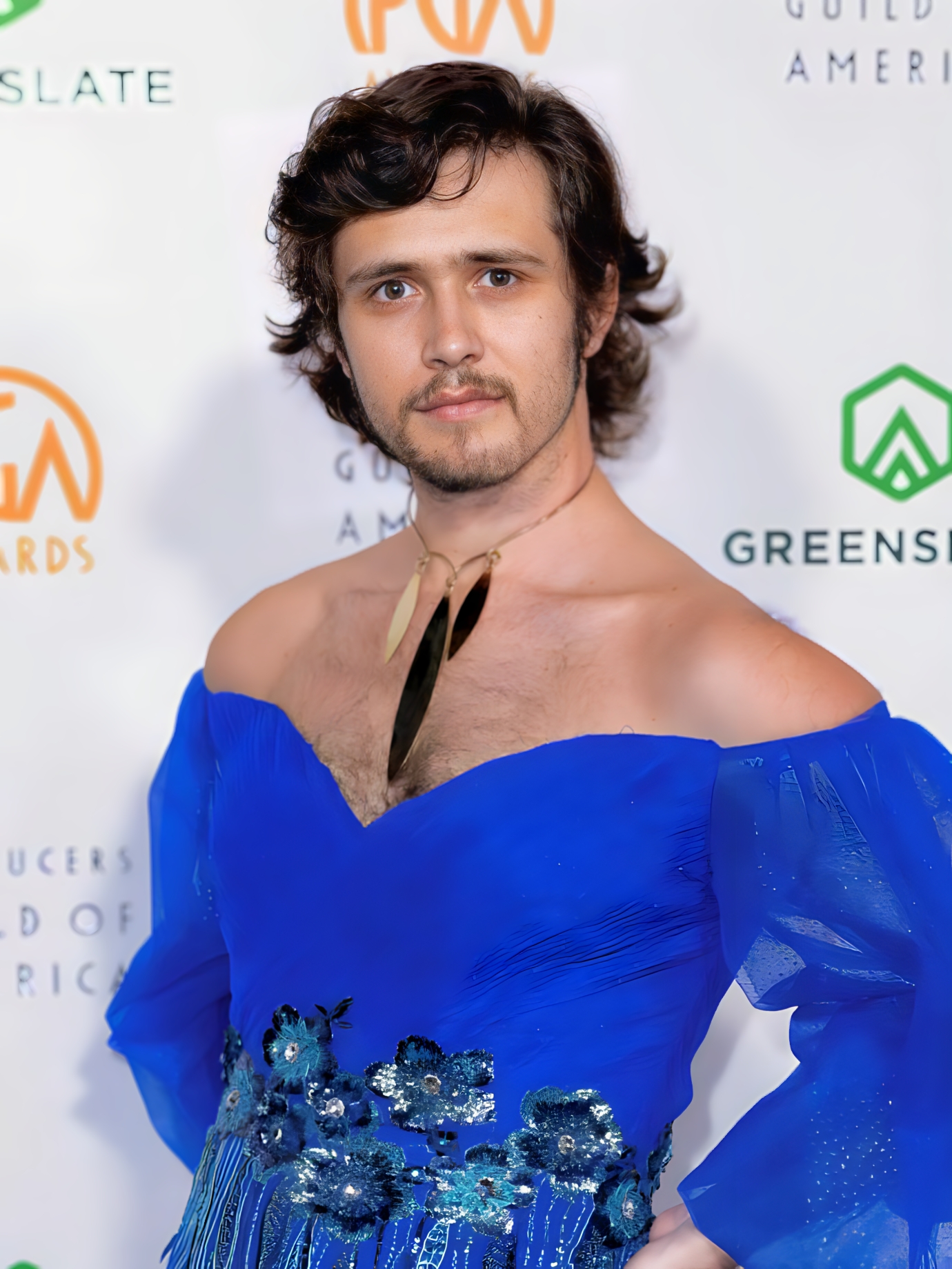 a person with shaggy brown hair wears a blue dress and poses in front of a step and repeat backdrop