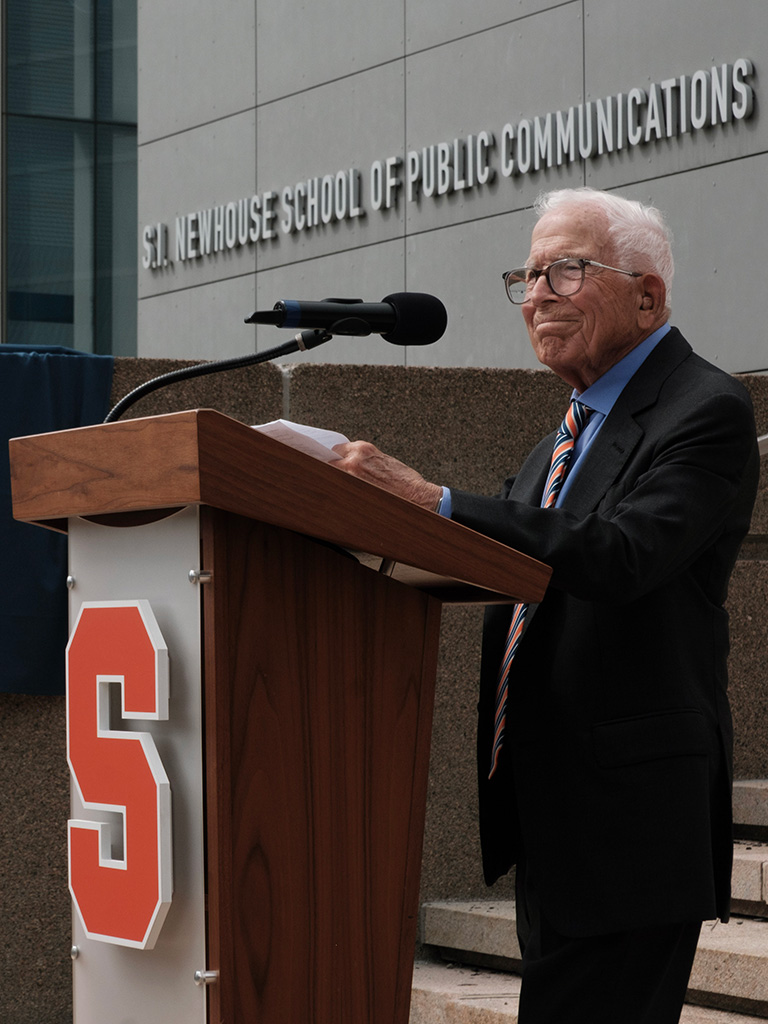 A man delivers remarks from a podium with a sign that reads "S.I. Newhouse School of Public Communications" in the background