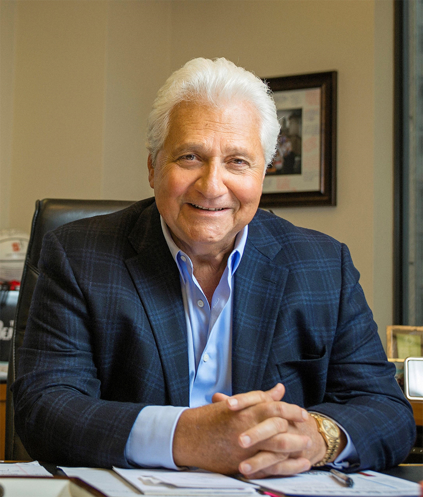 a person in a suit jacket and blue button down shirt sits at a desk with their hands folded