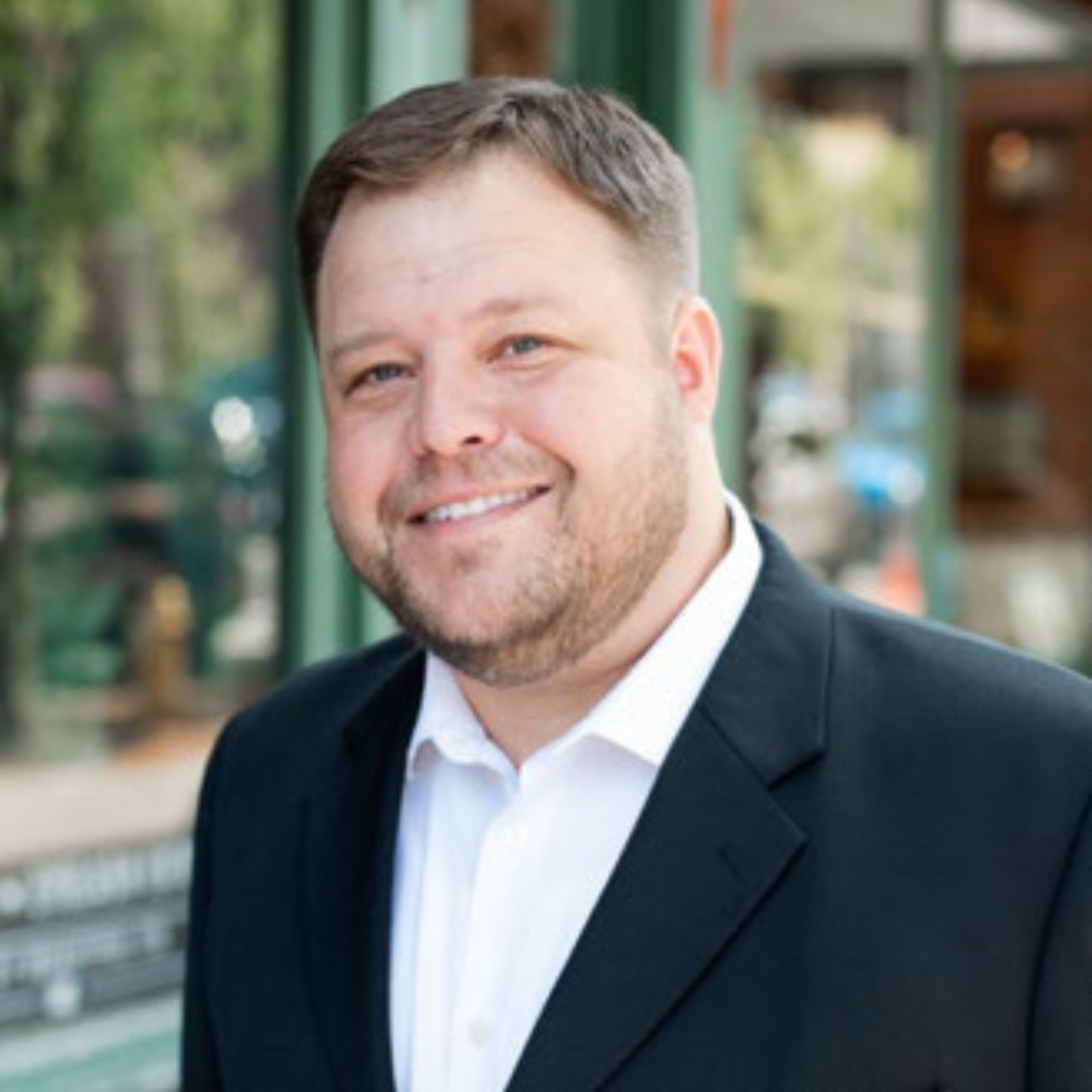  person with short light brown hair and facial hair smiles for the camera in a professional headshot. they wear a white shirt and black blazer