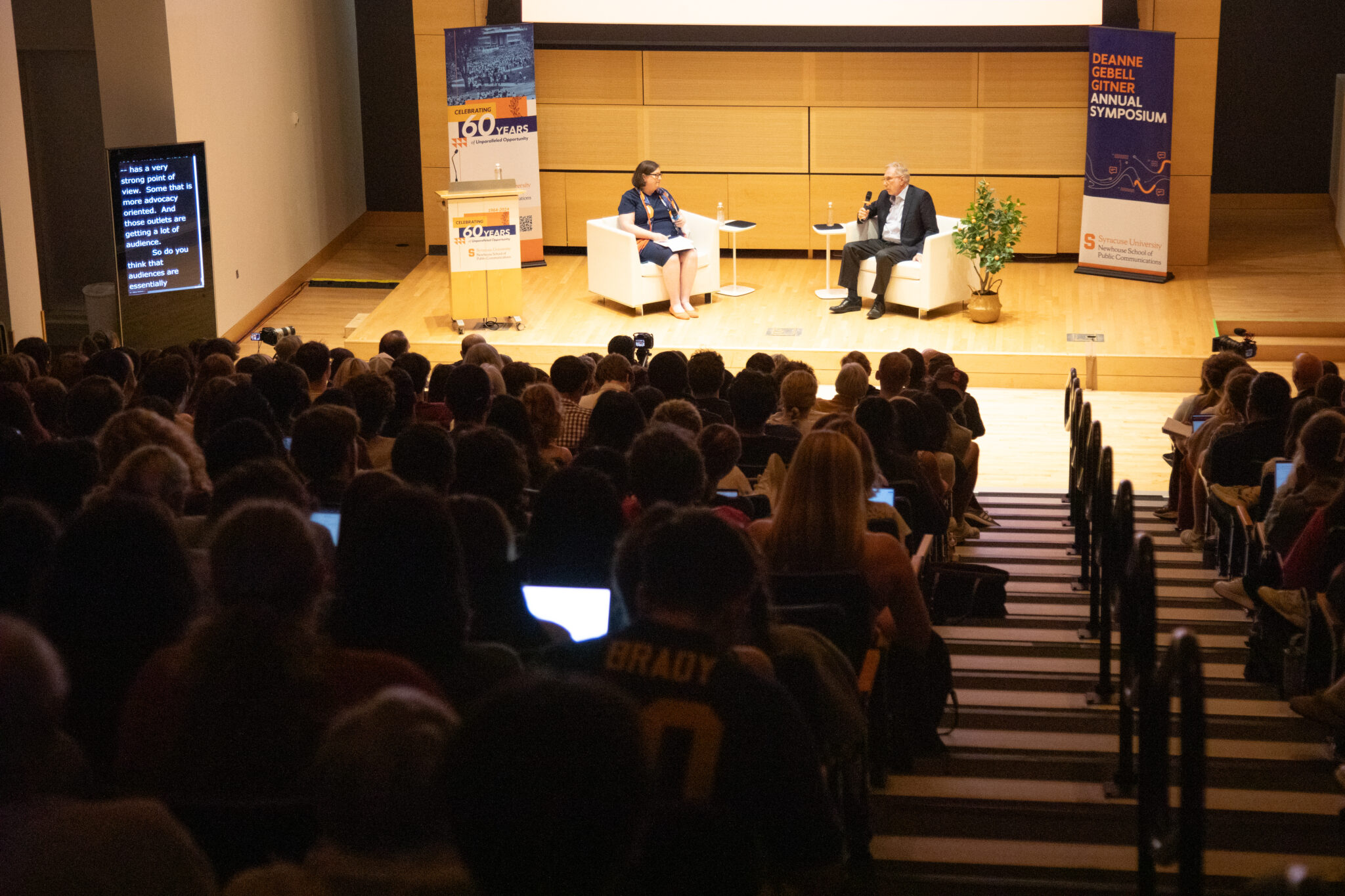 two people sit in chairs and talk in a packed auditorium